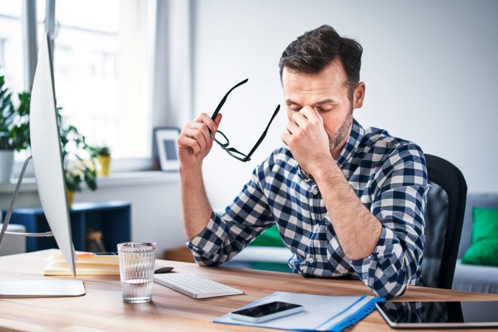 Man taking off his glasses looking stressed while sitting at a work station in his home